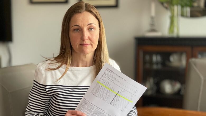 A white woman in a striped long-sleeved shirt sits are her kitchen table holding up a credit card statement with one line highlighted. 