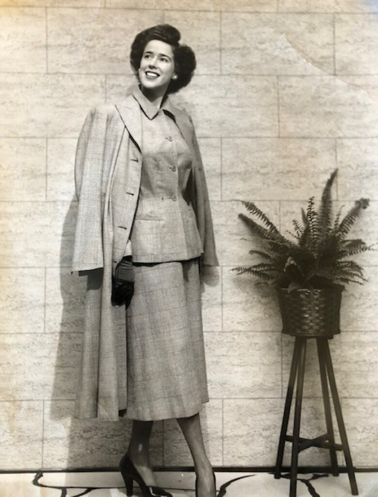 A monochrome photo of a woman in a cinched jacket and mid-calf length skirt wearing a cape.