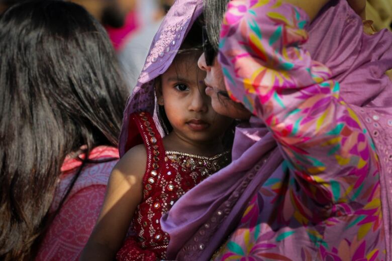 A mother holds a small girl, both wearing elaborate clothes on a sunny day.