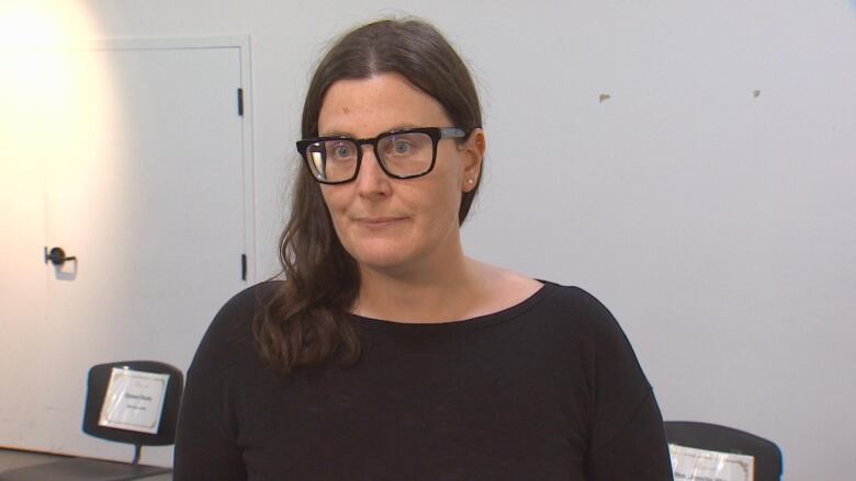 A woman in glasses looks at the camera in a white room.