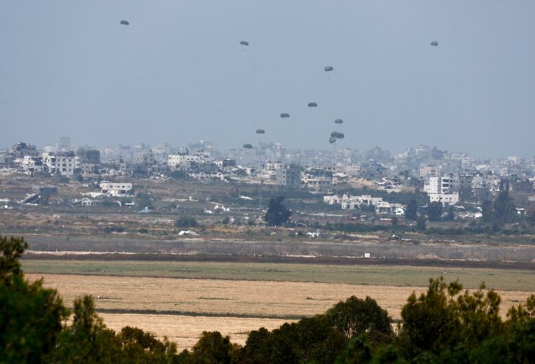 A shot from distance over a field shows a city with large balloons flying overhead.