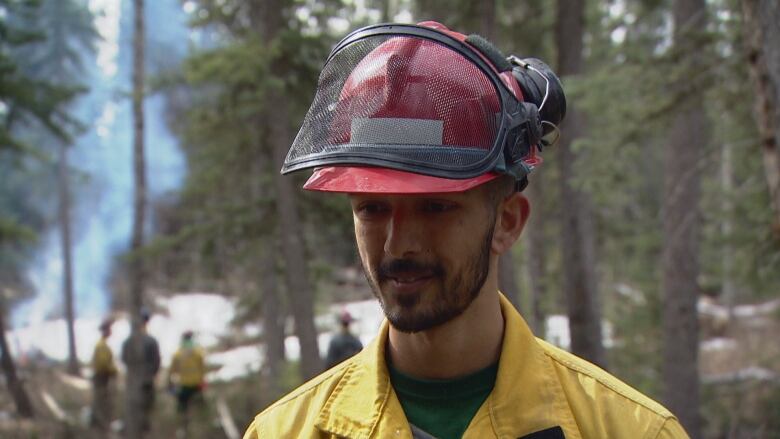 A man with a beard wearing a red helmet and Alberta Wildfire uniform stands in a forest.