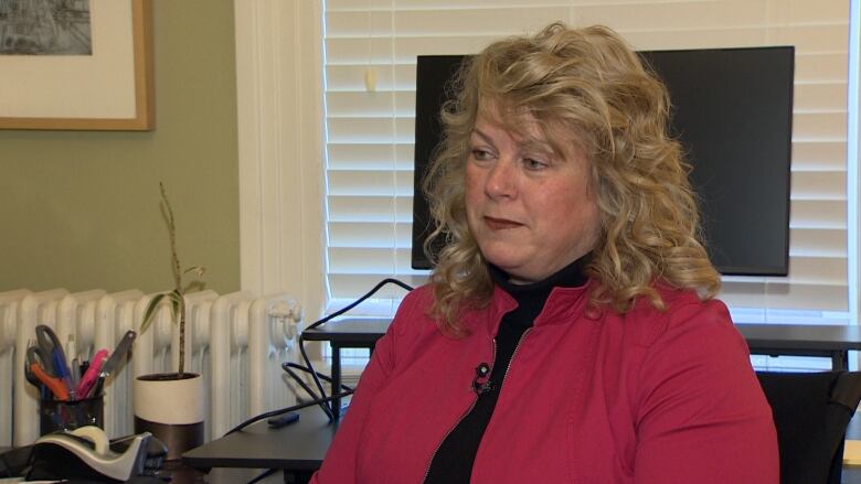 A woman with blonde, curly hair wears a red coat. She is sitting in her office in front of a computer screen.