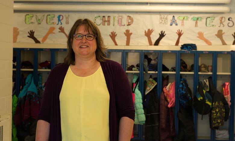 A woman in a yellow shirt with shoulder-length brown hair and glasses stands in a school by a banner that reads 'every child matters.'