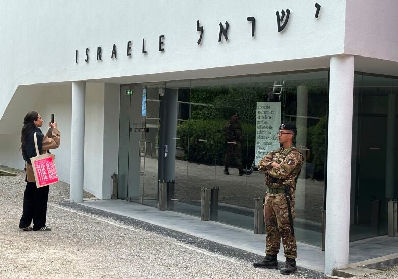 A woman takes a photo as a soldier wearing a hat and camouflage clothing stands nearby in front of a closed building.