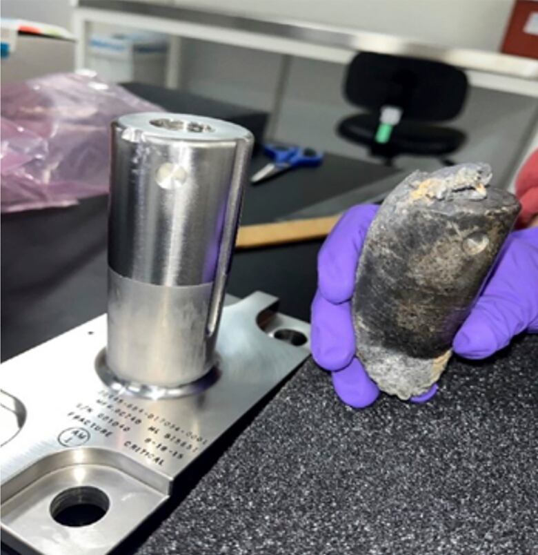 A gloved hand is seen holding a piece of degraded metal in a lab.