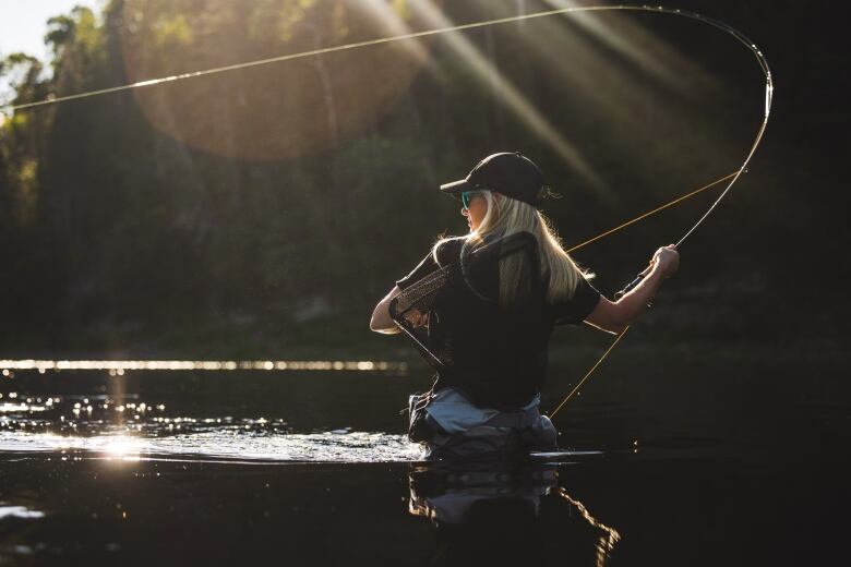 A blond woman stands in a river casting a fishing rod. 