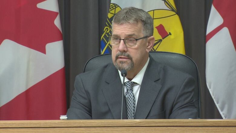 A man in a grey suot and glasses sits at a podium desk with flags behind him.