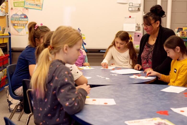 A teacher with black hair in a pony tail is surrounded by young students at two tables covered in bingo cards.