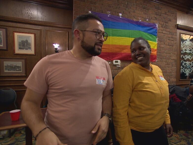 A man and woman dance beside each other at a bar in front of a rainbow flag.