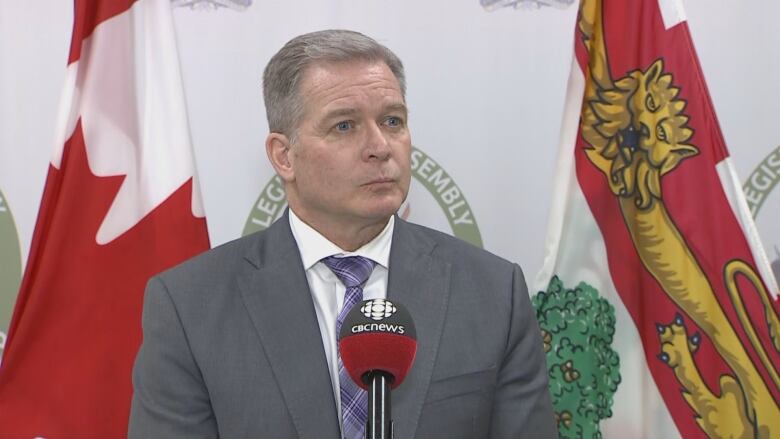 A man in a grey suit and purple tie stands in front of a microphone taking questions from reporters.