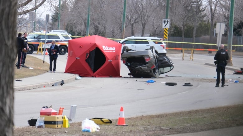 A red tent sits on the road beside an upside down vehicle, with car pieces scattered on the road and police officers standing nearby.