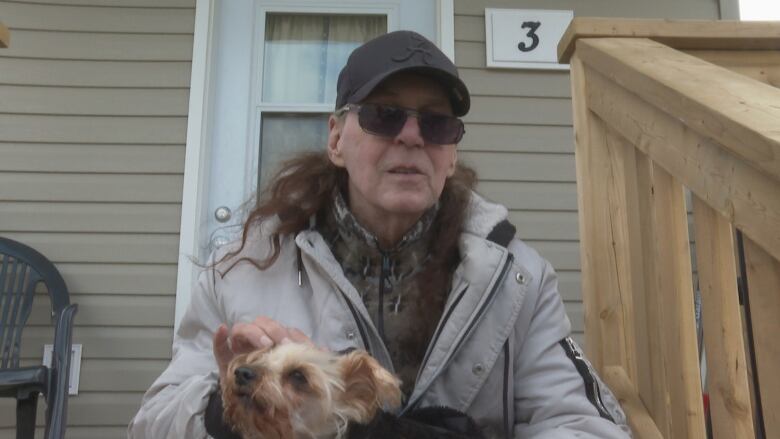 A man with long brown hair wearing clip-on sunglasses, a black baseball hat and a white coat over a camouflaged sweater sits deck stairs petting a small brown and black dog.