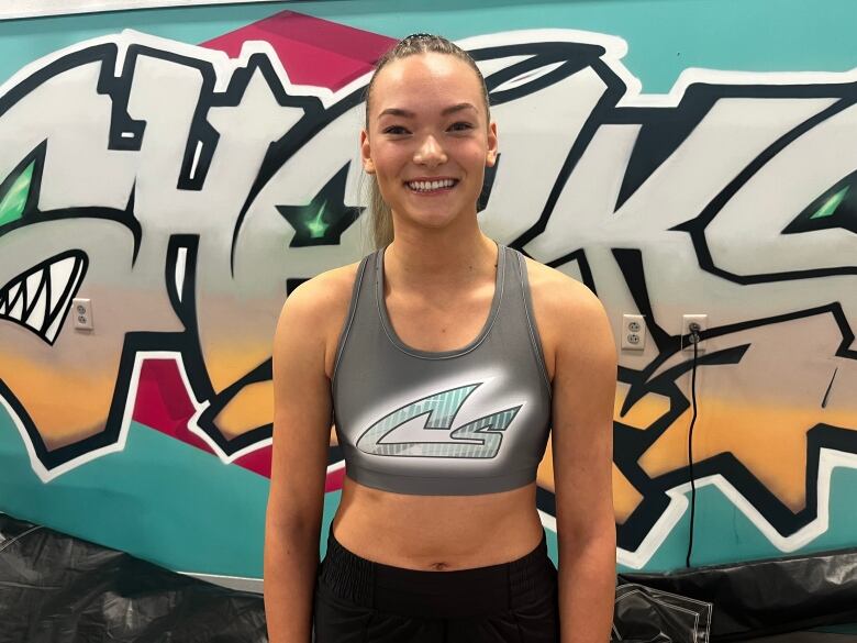 Dark haired woman in fitness clothing with her hair in pony tail stands in front of a Sharks logo. 