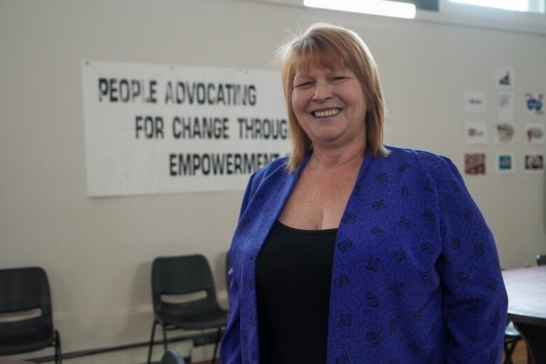 A person with red hair wearing a blue-purple blazer and black top stands inside a room, smiling.