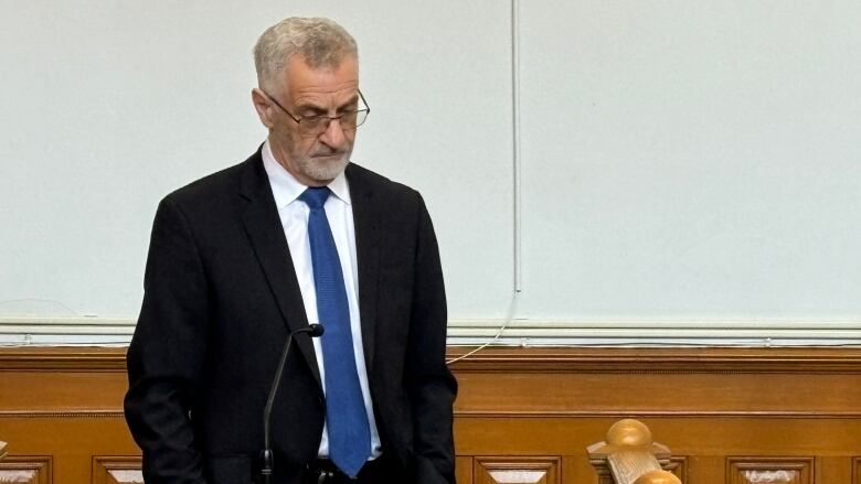 A man with gray hair and glasses is wearing a suit. His hands are in his pockets and he is looking down. 