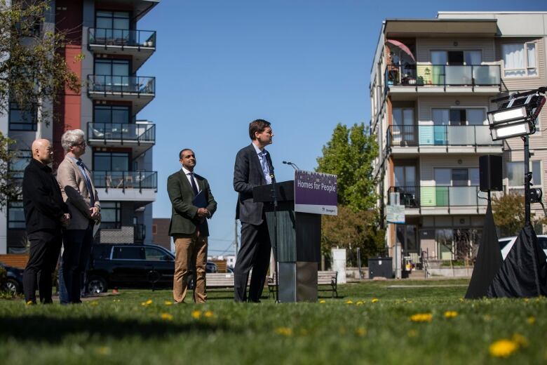 A wide shot of a man at a podium and microphone with multi-unit dwellings behind him.