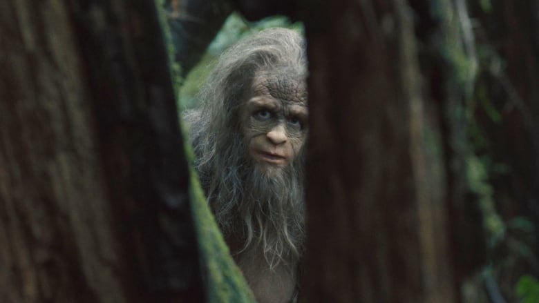 A man wearing a sasquatch costume gazes through a parting in the trees. 