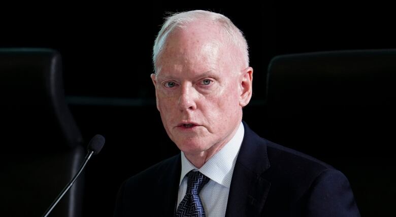 An older man wearing a blue tie and suit looks into the camera.