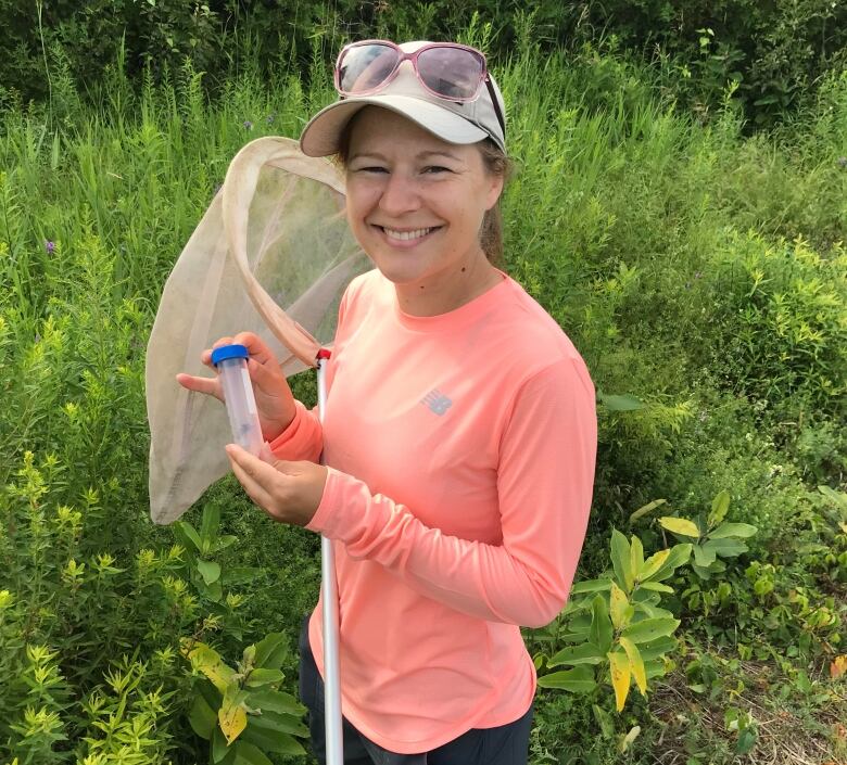 Dr. Sabrina Rondeau stands in front of foliage holding a net and tube for catching bees. 