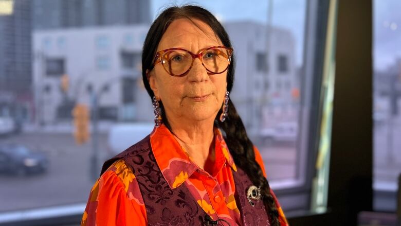 A woman wears glasses and a brightly-coloured shirt. She has long black hair in a braid over her shoulder.