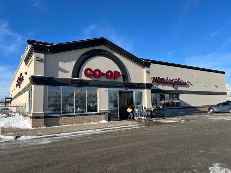 In the foreground is a large parking lot with a but of snow around the edges and one parked car on the right of the image. In the background is a beige and brown building with large windows and automatic sliding doors in the front with one shopper exiting the doors. On the upper left front of the store exterior there is a mounted sign that says 