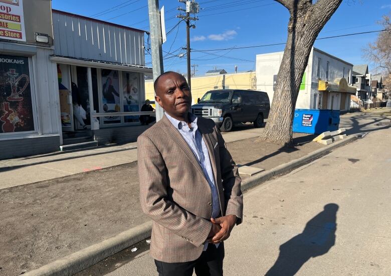 Man in suit stands on road with a shop and house in the background.