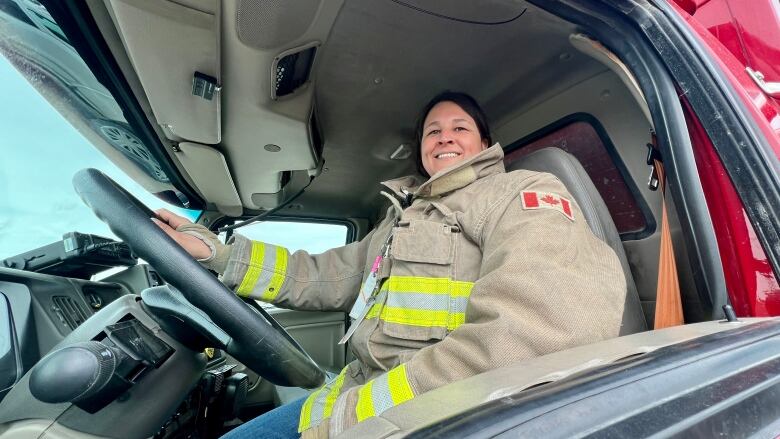 A woman in firefighter gear sits behind the wheel of a fire engine.