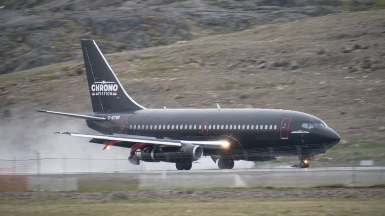 A black plane takes off from a runway with tundra in the background. 