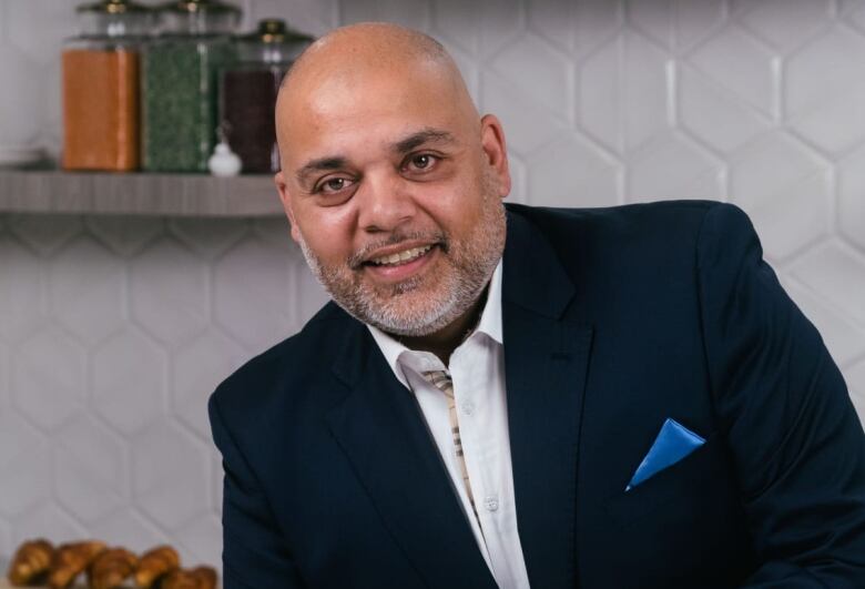 A bald man in a jacket and collared shirt leans on a counter.