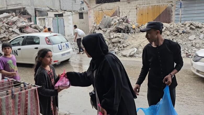 A man and woman hand bags of food to children in a street lined with rubble. A U.N. vehicle is in the background.