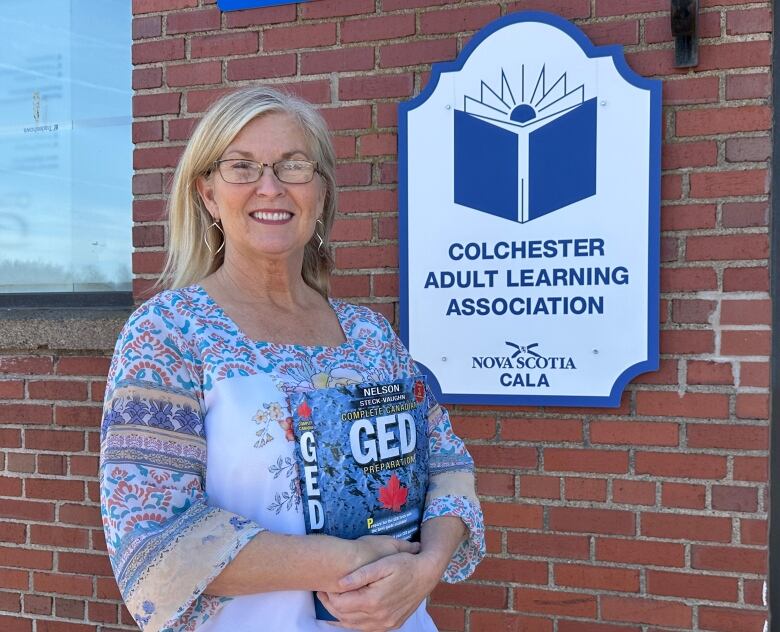 A woman wearing a floral blouse a with light wash jeans stands in front of a brick building with a sign that says Colchester Adult Learning Association. The woman is holding a large textbook that says GED. 