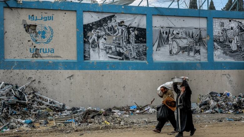 Two people, carrying items on their heads, walk past a bullet-riddle concrete wall with three black and white images painted along the the top and, in the left corner, a blue United Nations logo with the Arabic letters above and the word UNRWA below. 