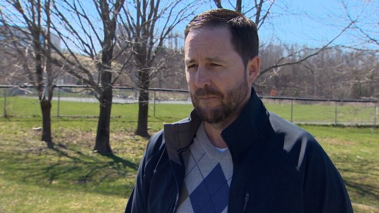 A man stands outside in front of a ball field. He has brown hair and a brown beard. He wears a black jacket and a grey and blue sweater underneath. 