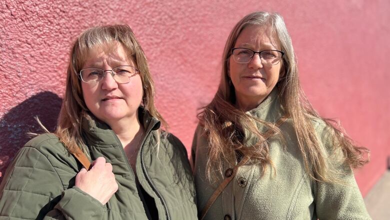 Two women stand next to each other, both wearing green coats and glasses.