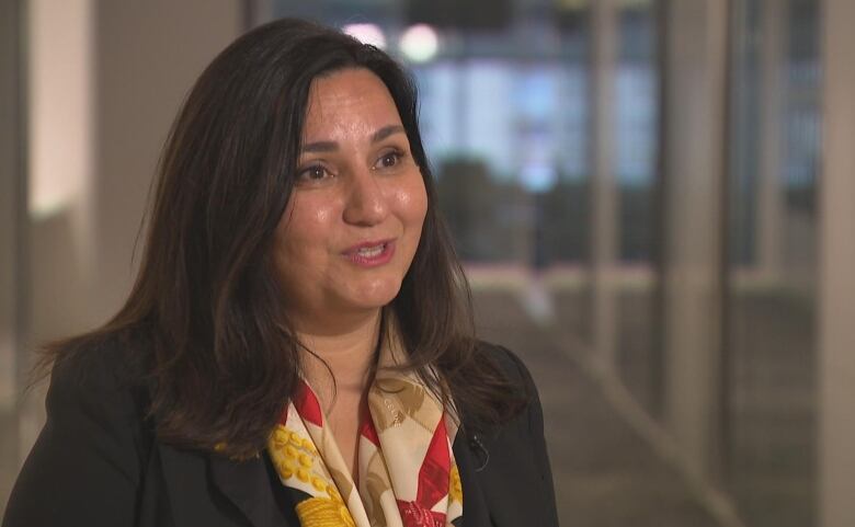 A woman in an orange and red scarf looks to the side of the camera while being interviewed.