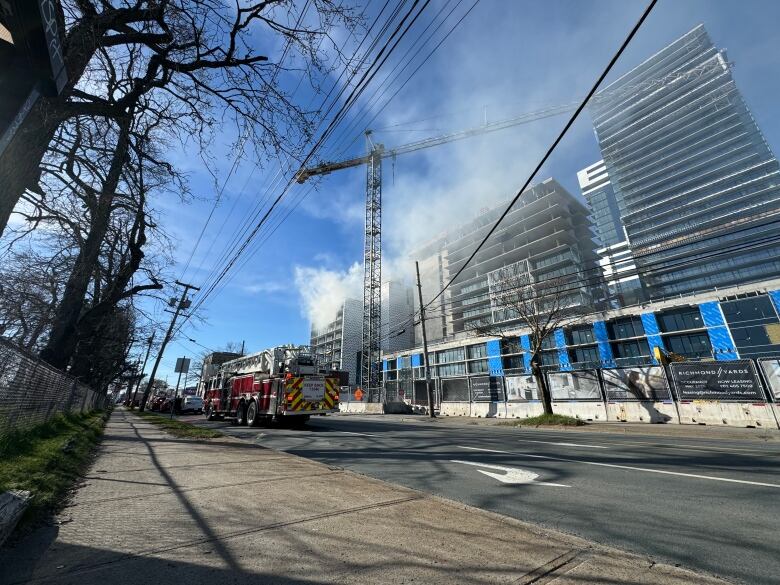 A fire truck is seen driving down a city street, with an under construction building in the background and white plumes of smoke coming from the top.