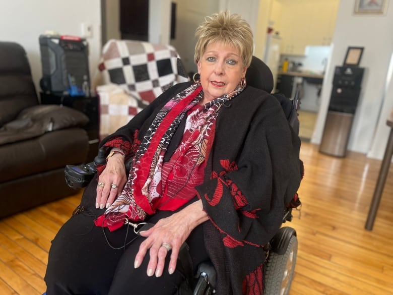 A woman in a wheelchair looks at the camera, she is in her living room, her kitchen is seen in the background out-of-focus.