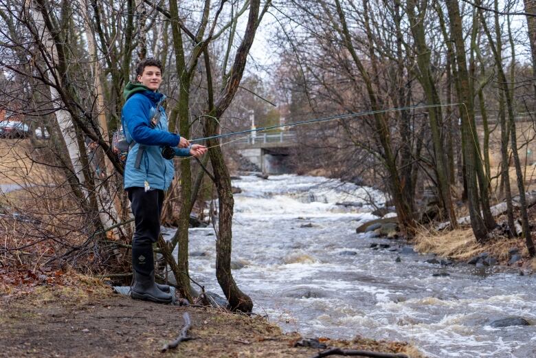 A young man stands on the bank and casts his fishing rod into the water. 