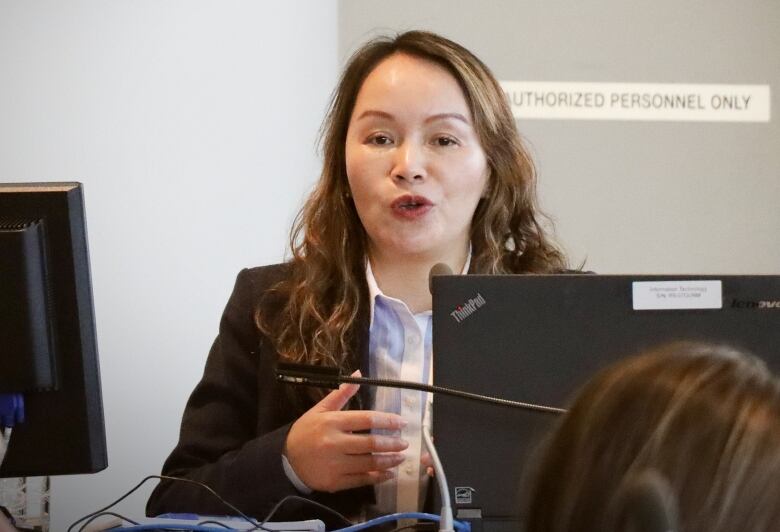 A woman with a dark blazer and light coloured blouse speaks in front of a microphone and computer screen.