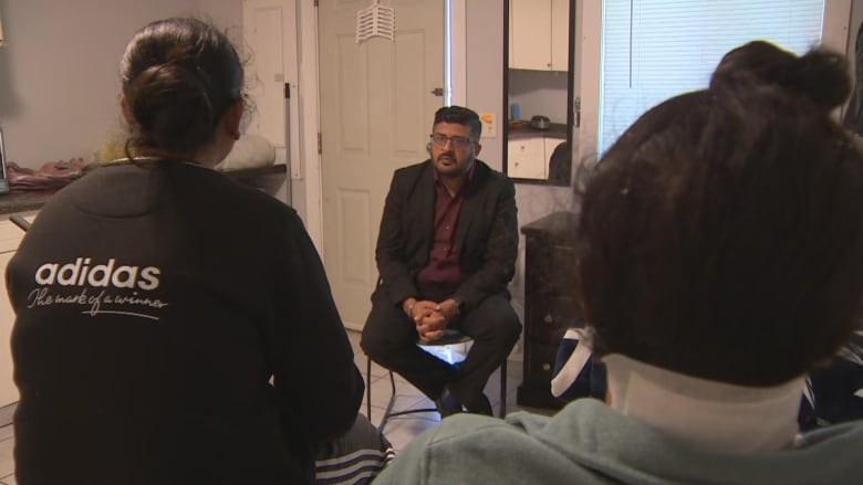 A South Asian man listens to two people with their backs to the camera. The person on the right of the image has bandages on their neck.