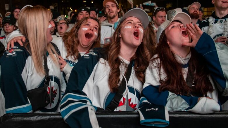 Girls in Winnipeg Jets jerseys cheer amid a sea of fans also wearing white.