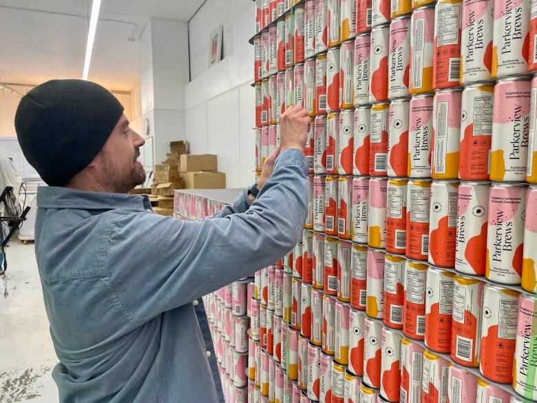 A man wearing a blue shirt and sporting a beanie moves towards a wall stacked with cans.