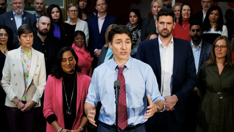 Prime Minister Justin Trudeau stands at a microphone holding up his hand. A crowd stands behind him as he answers a media question. 