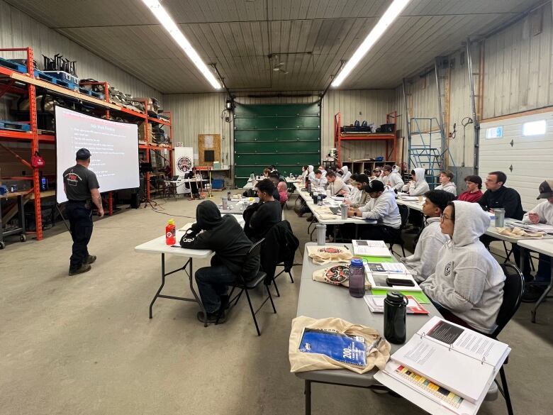 A classroom set up inside a garage. Students in grey hoodies are looking at an instructor with a screen. 