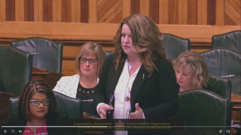 A woman with long hair is standing and speaking in a room with other women sitting around here.