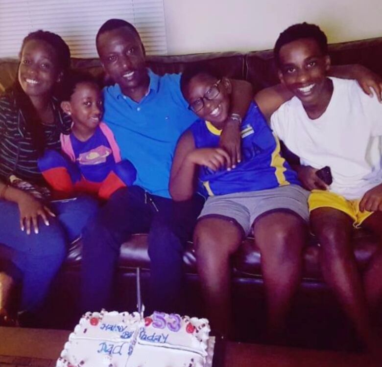 A family of five sits on a couch with a birthday cake on the living room table. 