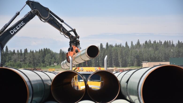 In front to blue Sky and fir trees the mechanical arm of a large machine swings a pipe onto a stack of pipes at a construction yard in Saskatchewan. 