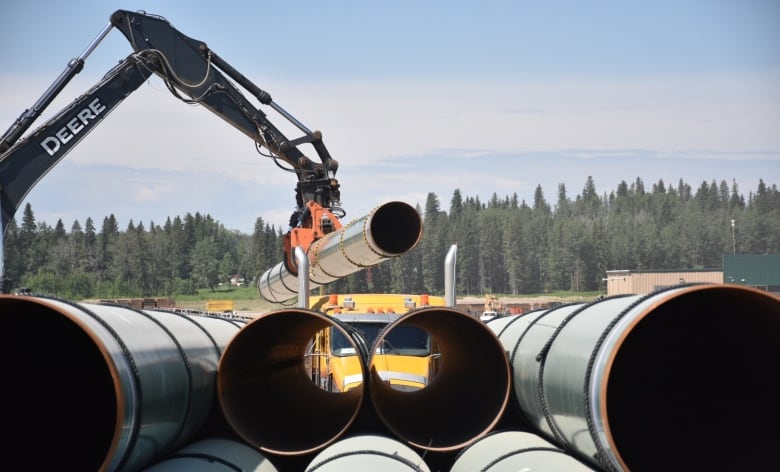 In front to blue Sky and fir trees the mechanical arm of a large machine swings a pipe onto a stack of pipes at a construction yard in Saskatchewan. 