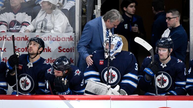 A man in a suit talks to a goaltender seated on the team's bench.